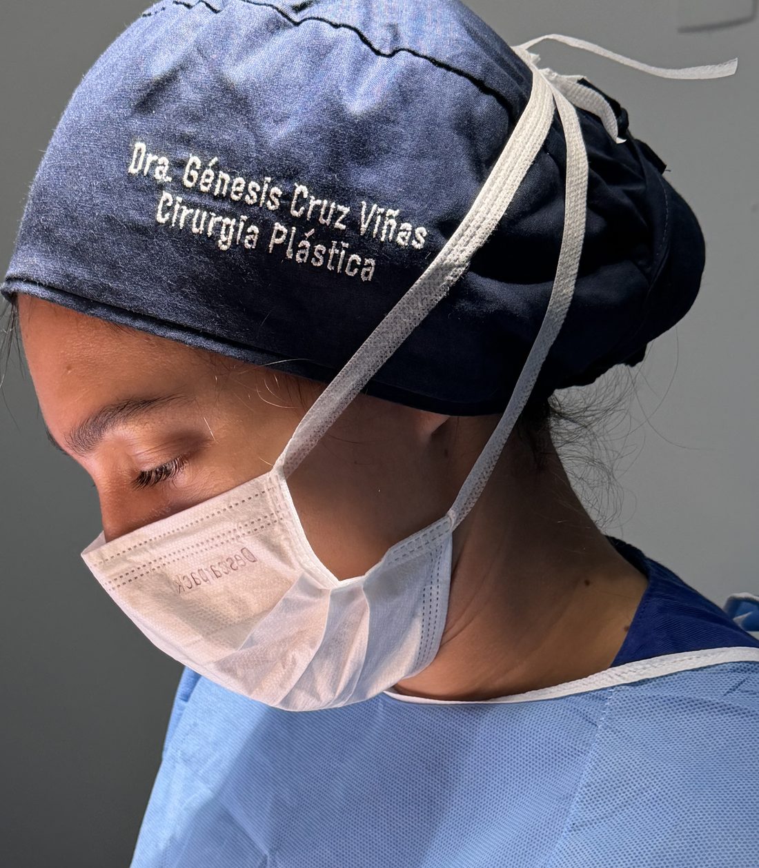 La Dra. Génesis Cruz Viñas en quirófano, con gorro quirúrgico bordado con su nombre y la especialidad de Cirurgia Plástica.