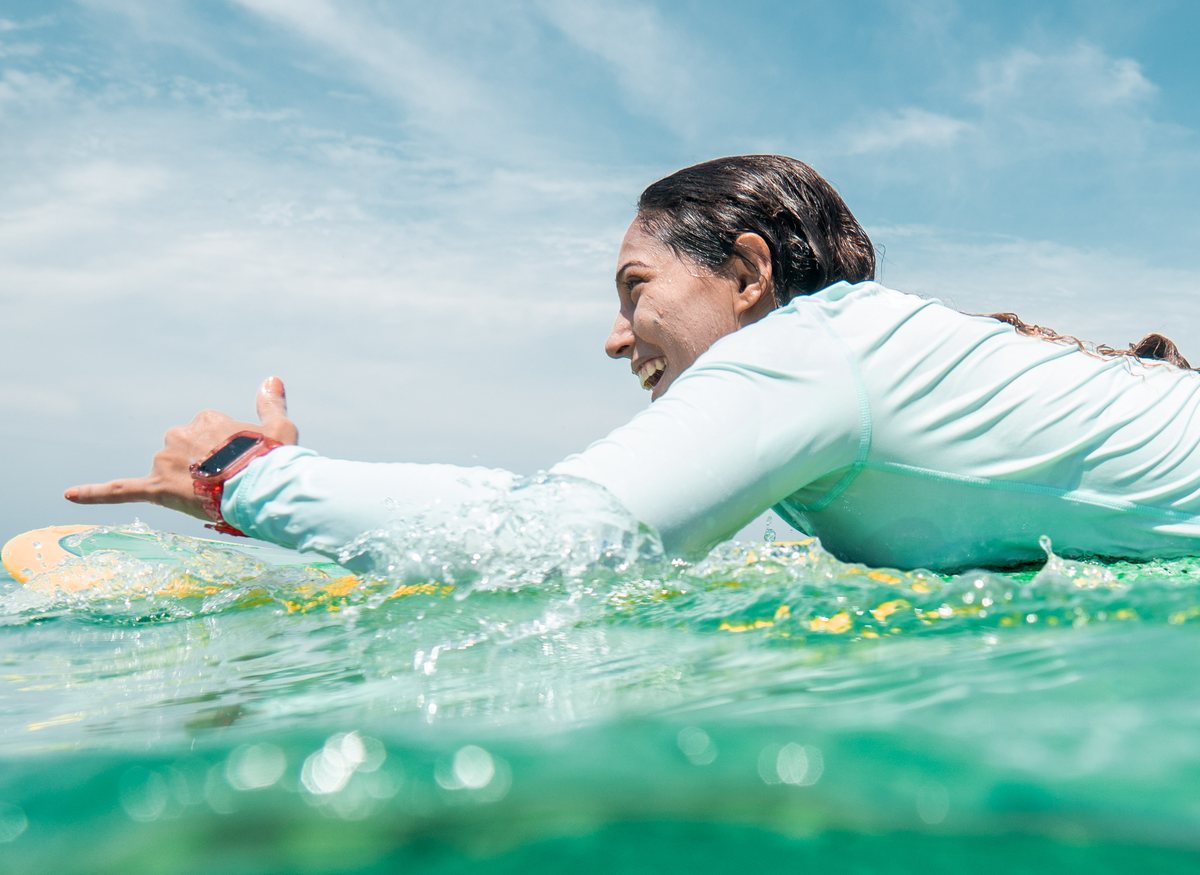 La Dra. Génesis Cruz Viñas remando sobre una tabla de surf, riendo, con licra de manga larga.